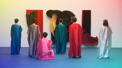 Film still from 'Clubhuis is DEAD' (2015) by Jon Beaton. Six people in colourful togas stand in an exhibition space looking at an artwork consisting of colourful fabrics that echoes their appearance.